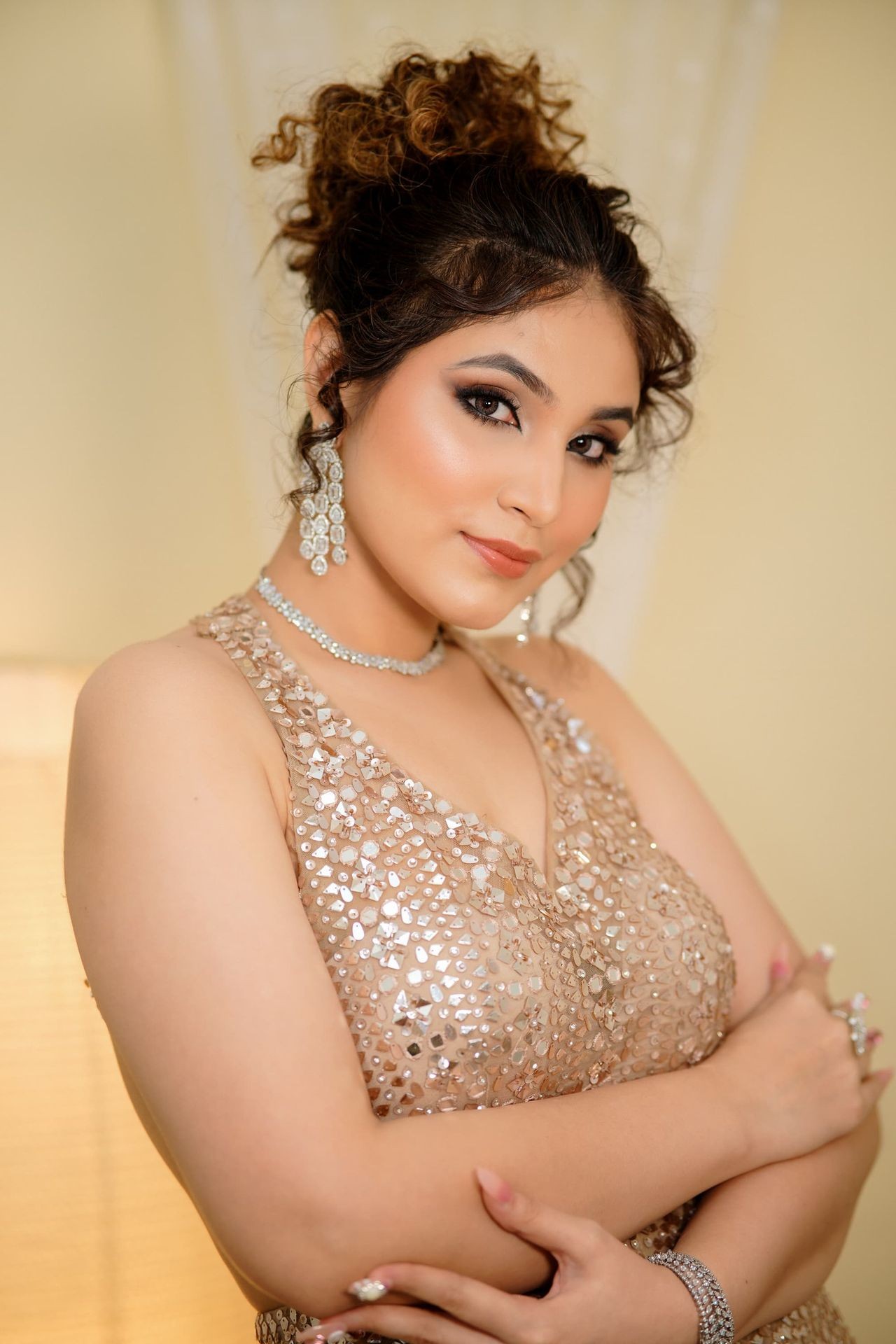 Woman with curly updo wearing a beaded dress and sparkling jewelry, posing with crossed arms.